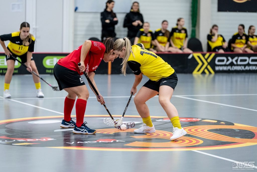 Divas florbolistes spēles sākumā centrā cīnās par bumbiņu iemetienā – viena sarkanā, otra dzelteni melnā formā. Fonā redzamas komandas biedrenes un rezervistu soliņš sporta zālē.