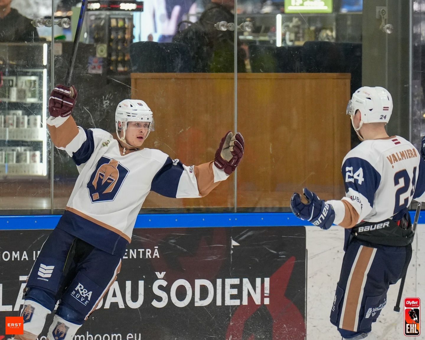 Divi hokejisti Valmieras formās svin vārtu guvumu pie laukuma apmales. Viens spēlētājs ar paceltām rokām slido pret komandas biedru ar 24. numuru. Fonā redzams stikla norobežojums un reklāmas laukuma malā.