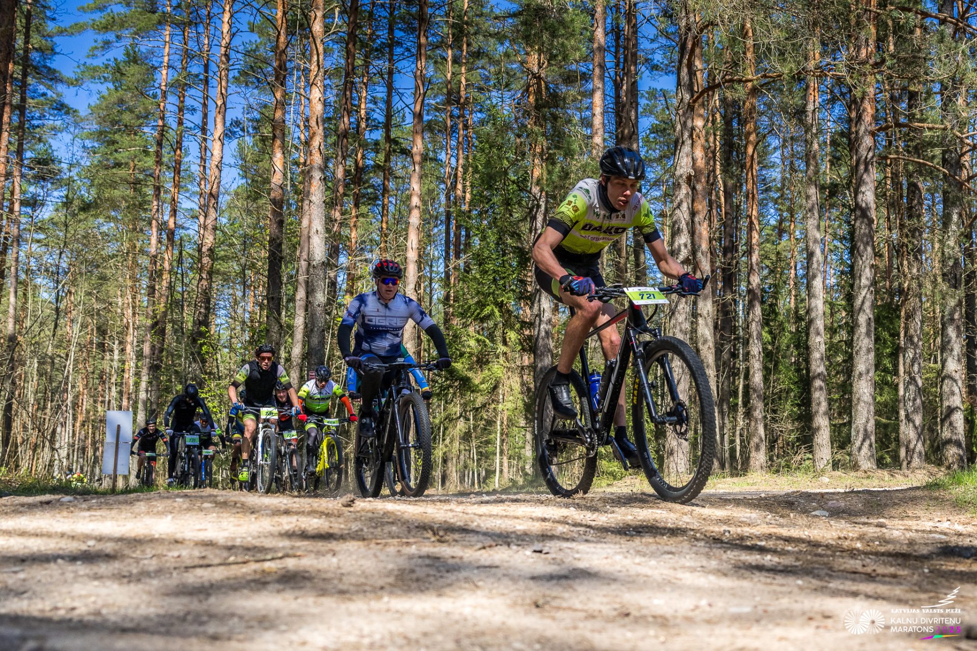 Kalnu divriteņu maratona dalībnieki brauc pa smilšainu meža ceļu priežu ieskautā trasē. Priekšplānā redzami vairāki riteņbraucēji, no kuriem viens izvirzījies vadībā, bet fonā aiz viņa seko pārējā grupa saulainā pavasara dienā.