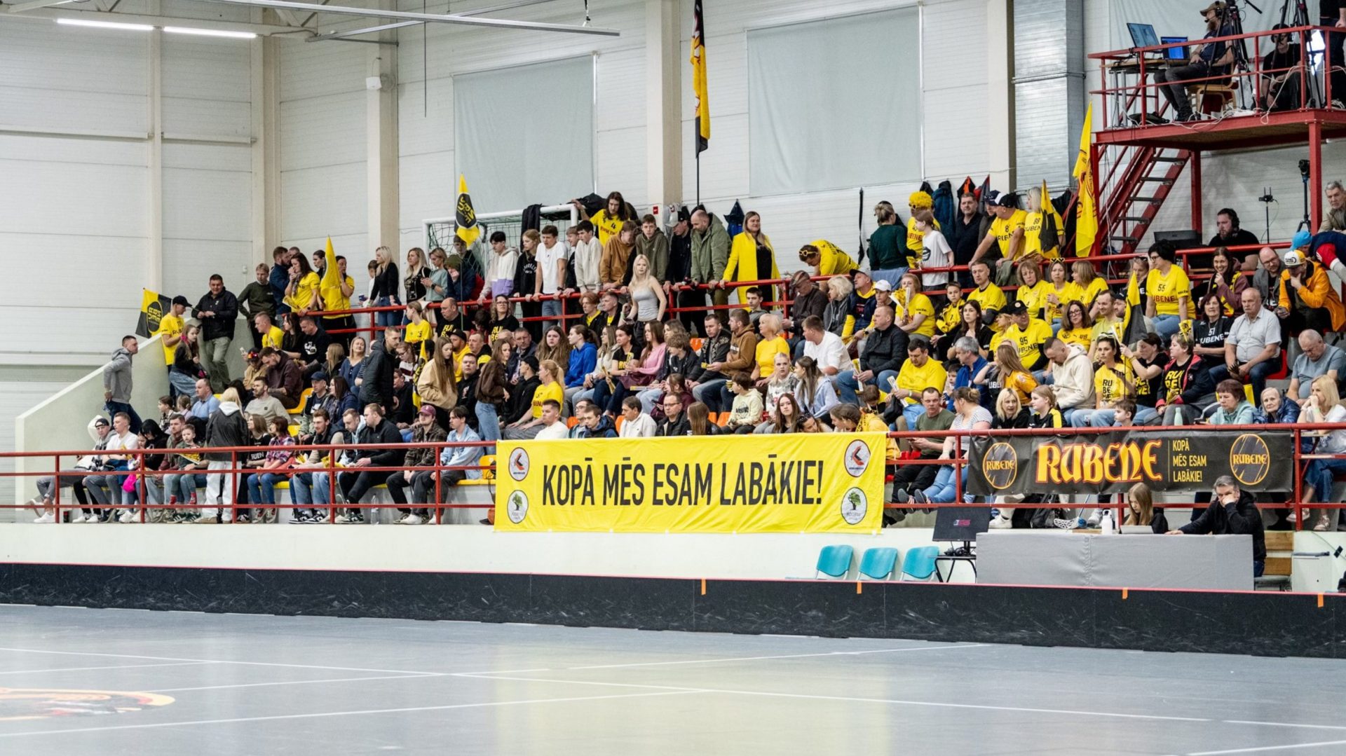Plašas tribīnes sporta hallē, kur pulcējušies daudzi līdzjutēji dzeltenos “Rubene” kreklos. Priekšplānā redzams liels dzeltens plakāts ar uzrakstu “Kopā mēs esam labākie!”, bet labajā pusē vēl vairāki “Rubene” fanu baneri un karogi.