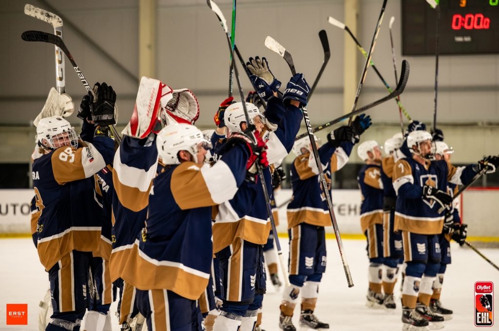 Hokeja komandas Valmiera spēlētāji pēc spēles stāv uz ledus ar paceltām nūjām un rokām, sveicot līdzjutējus. Spēlētāji ir tumši zilās un zeltītās formās, bet fonā redzama halles siena un rezultāta tablo.