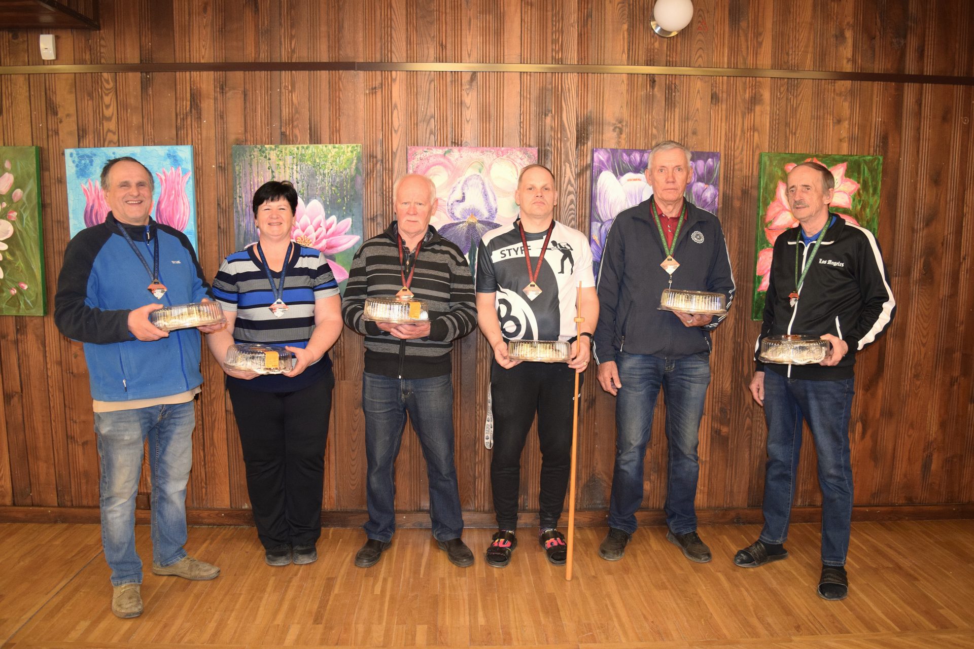 Seši novusa turnīra dalībnieki ar medaļām kaklā stāv rindā pie koka sienas ar ziedu gleznām un rokās tur balvās saņemtas tortes. Viens no vīriešiem stāv ar atbalsta spieķi.