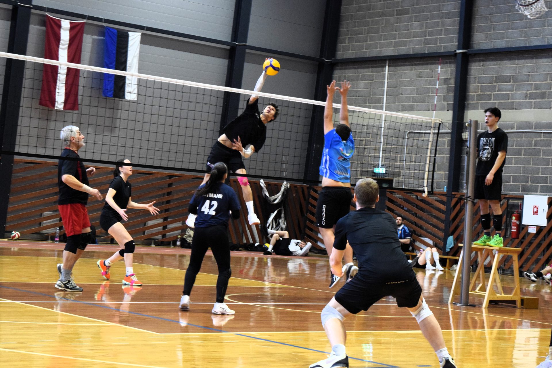 Volejbola spēles epizode sporta hallē – melnās formas spēlētājs lec pie tīkla un sit bumbu pāri pretinieka blokam, kamēr pārējie spēlētāji gatavojas nākamajai izspēlei. Fonā redzami Latvijas un Igaunijas karogi.