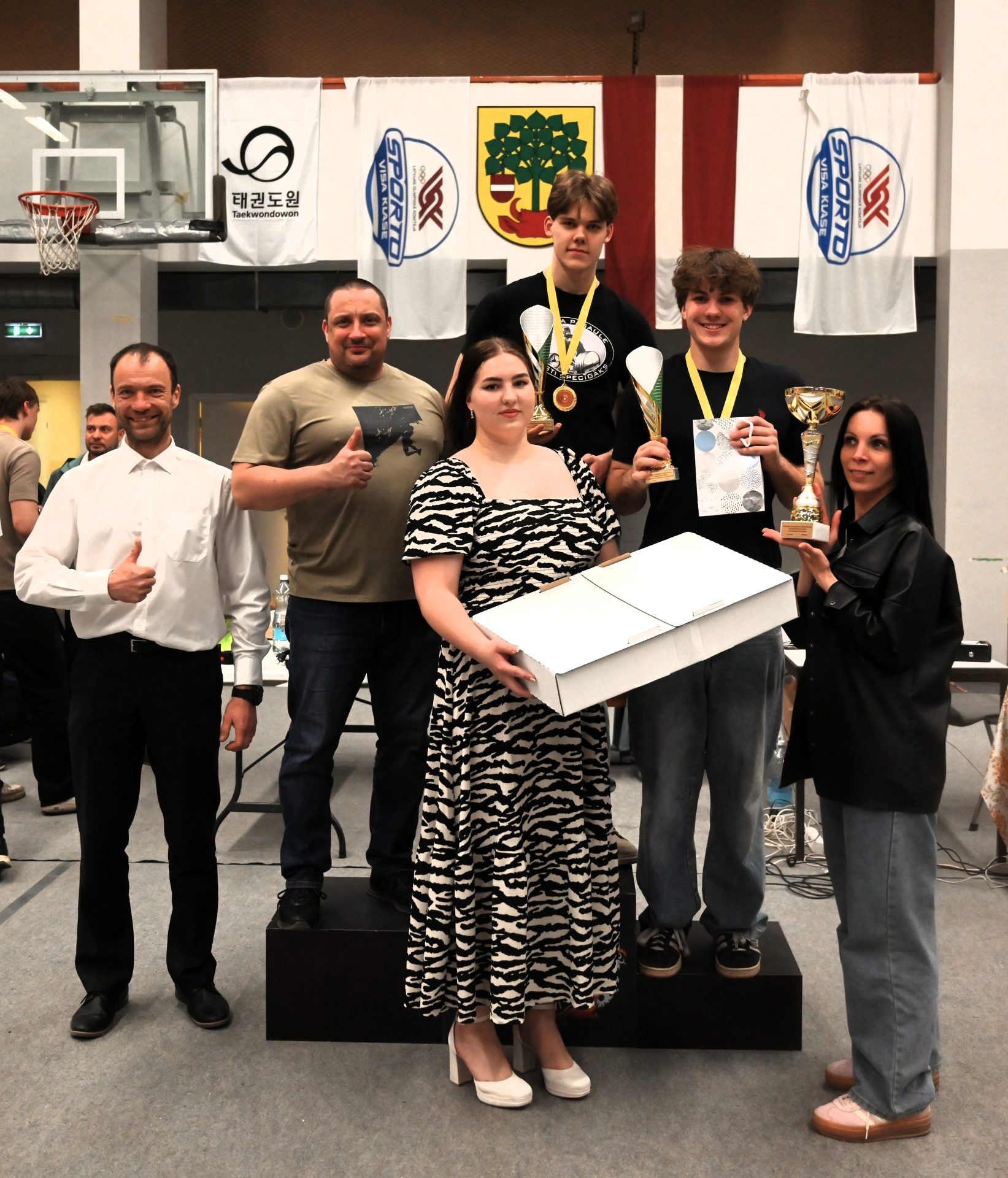 Apbalvošanas brīdis sporta zālē – uz pjedestāla un tā priekšā seši cilvēki ar kausiem, medaļām un balvām. Divi jaunieši ar medaļām tur kausus, priekšplānā sieviete zebras raksta kleitā tur lielu baltu balvu kasti, bet labajā pusē sieviete demonstrē uzvarētāju kausu. Fonā redzami sporta un Latvijas karogi.