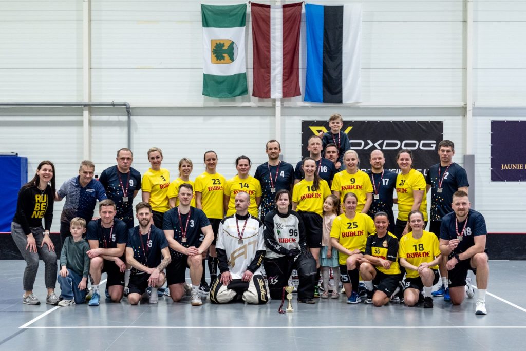 Grupas foto sporta zālē pēc florbola turnīra vai apbalvošanas. Attēlā redzami pieaugušie un bērni, daļa ģērbušies dzeltenos “Rubene” kreklos, citi tumšos sporta formas tērpos, vairākiem kaklā medaļas. Priekšplānā novietots kauss, bet fonā pie sienas redzami karogi un sporta zāles baneri.